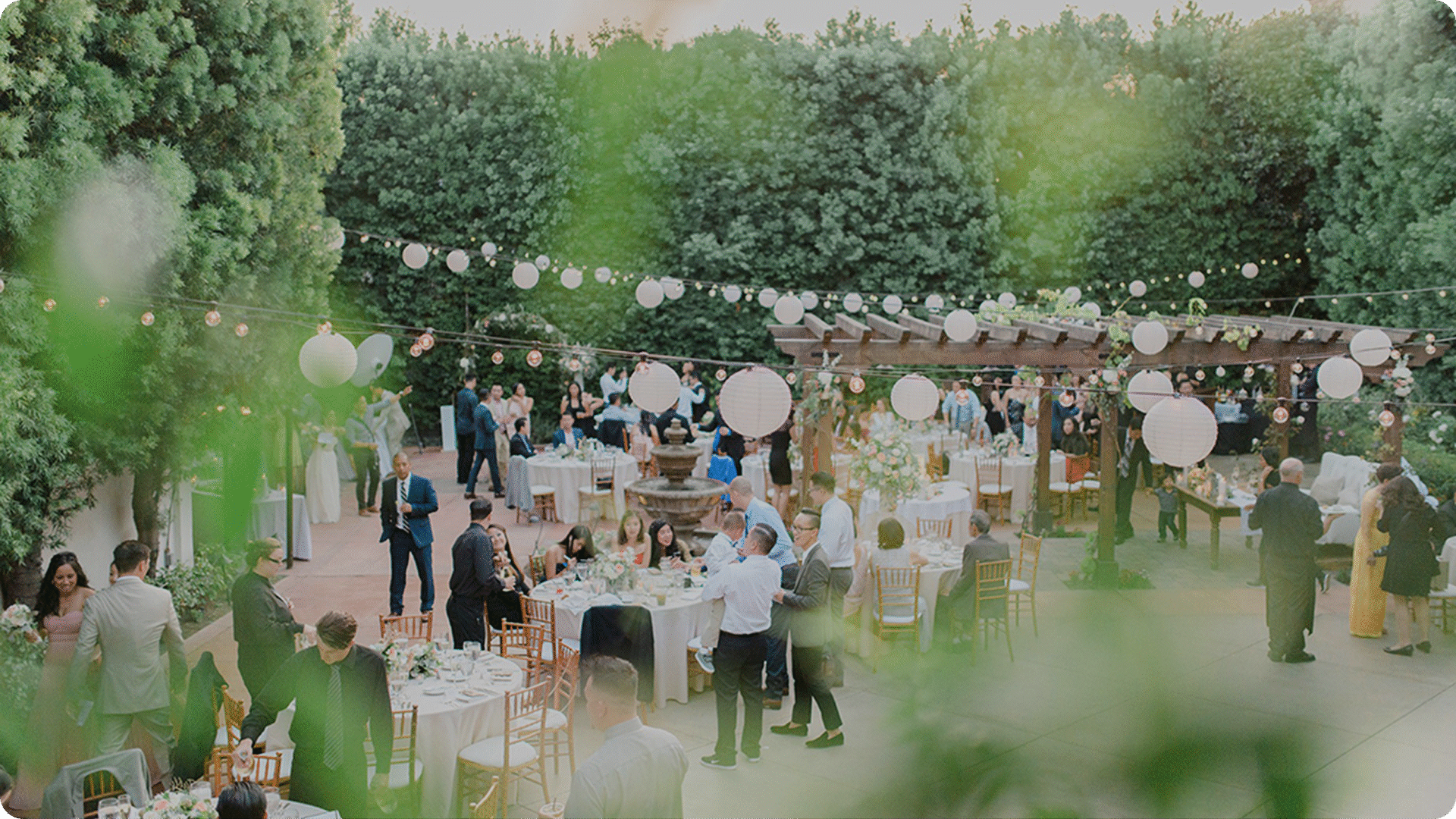 Outdoor wedding reception with guests mingling at decorated tables under string lights and paper lanterns. Greenery surrounds the festive courtyard, creating a warm, inviting atmosphere.