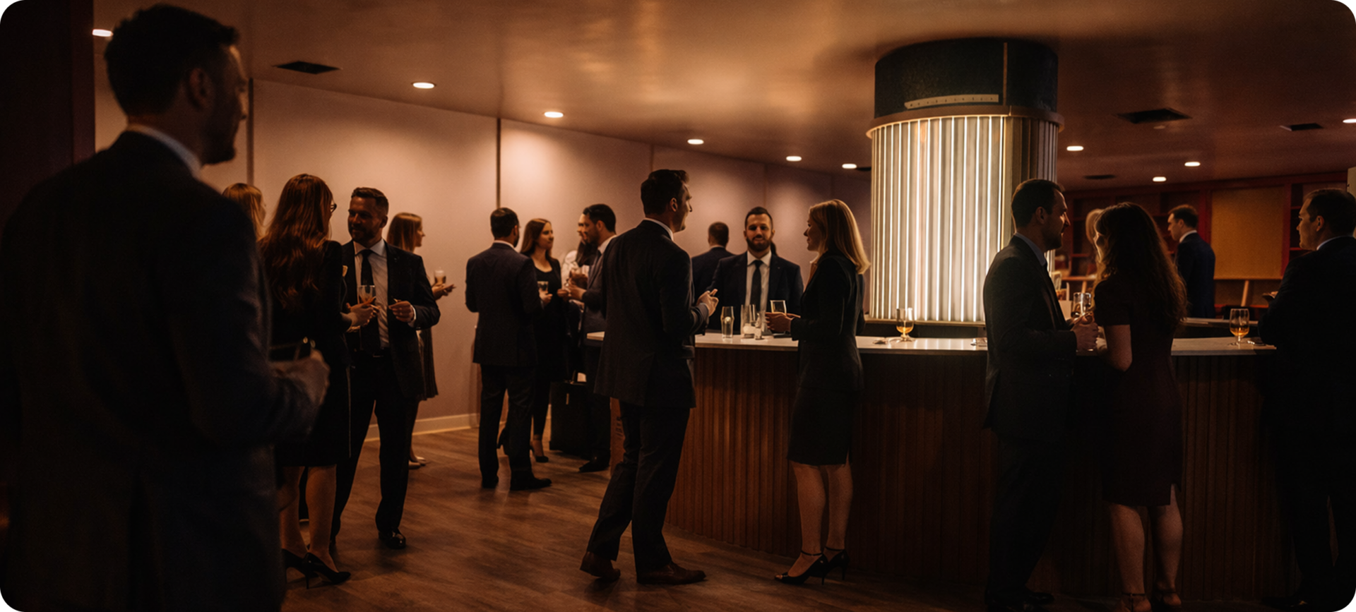 People in formal attire socialize and hold drinks around a modern, well-lit bar in a dimly lit room with warm tones.
