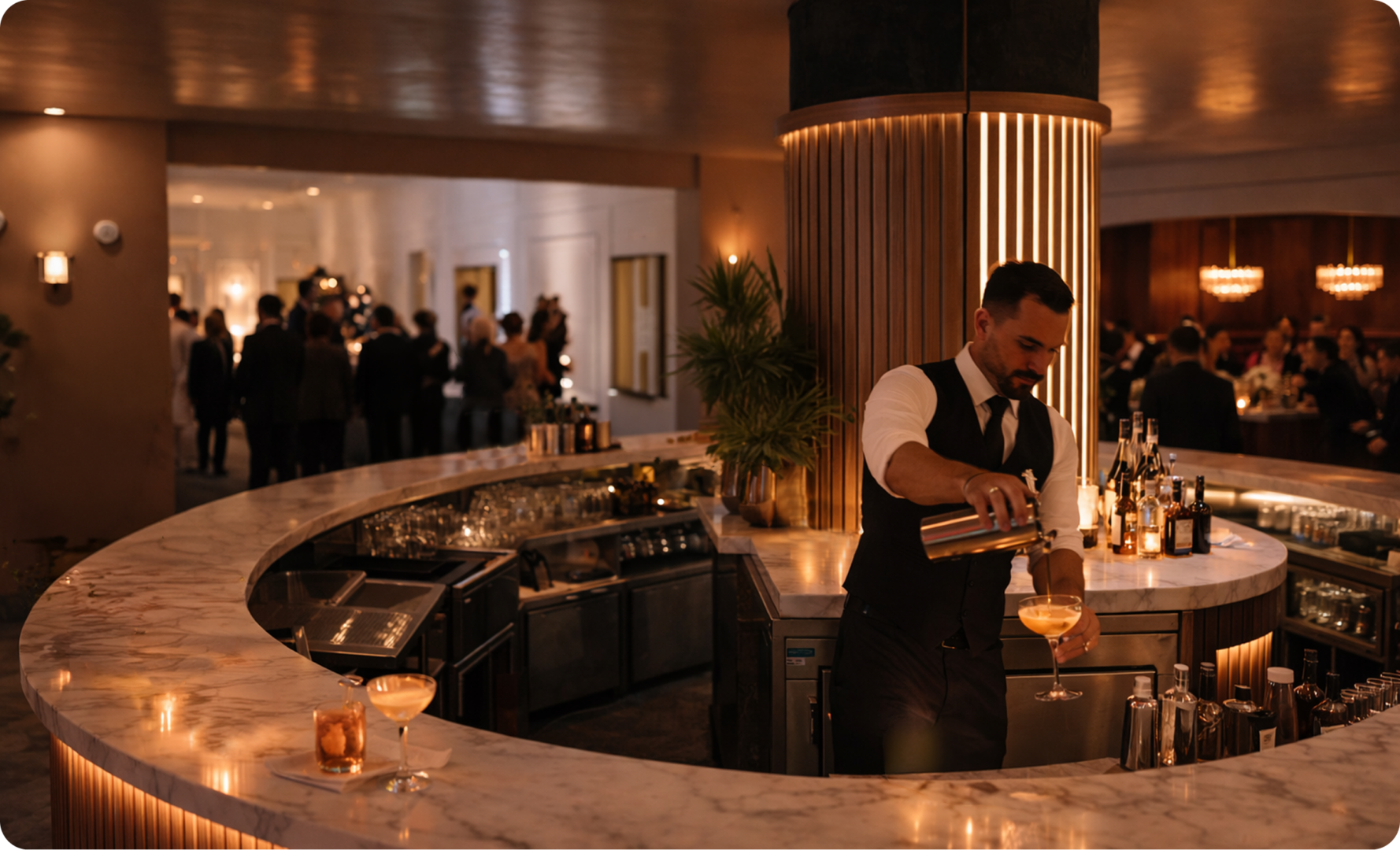 A bartender in a vest pours a cocktail behind a curved marble bar, with people socializing in a warmly lit background.