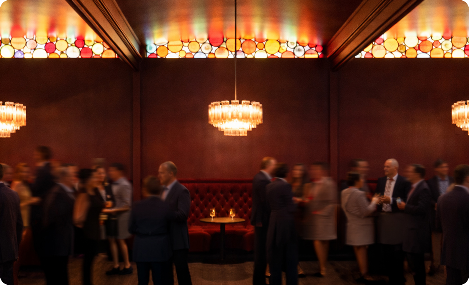 People in formal attire gather and converse in a dimly lit lounge with red booths, chandeliers, and stained-glass panels near the ceiling.