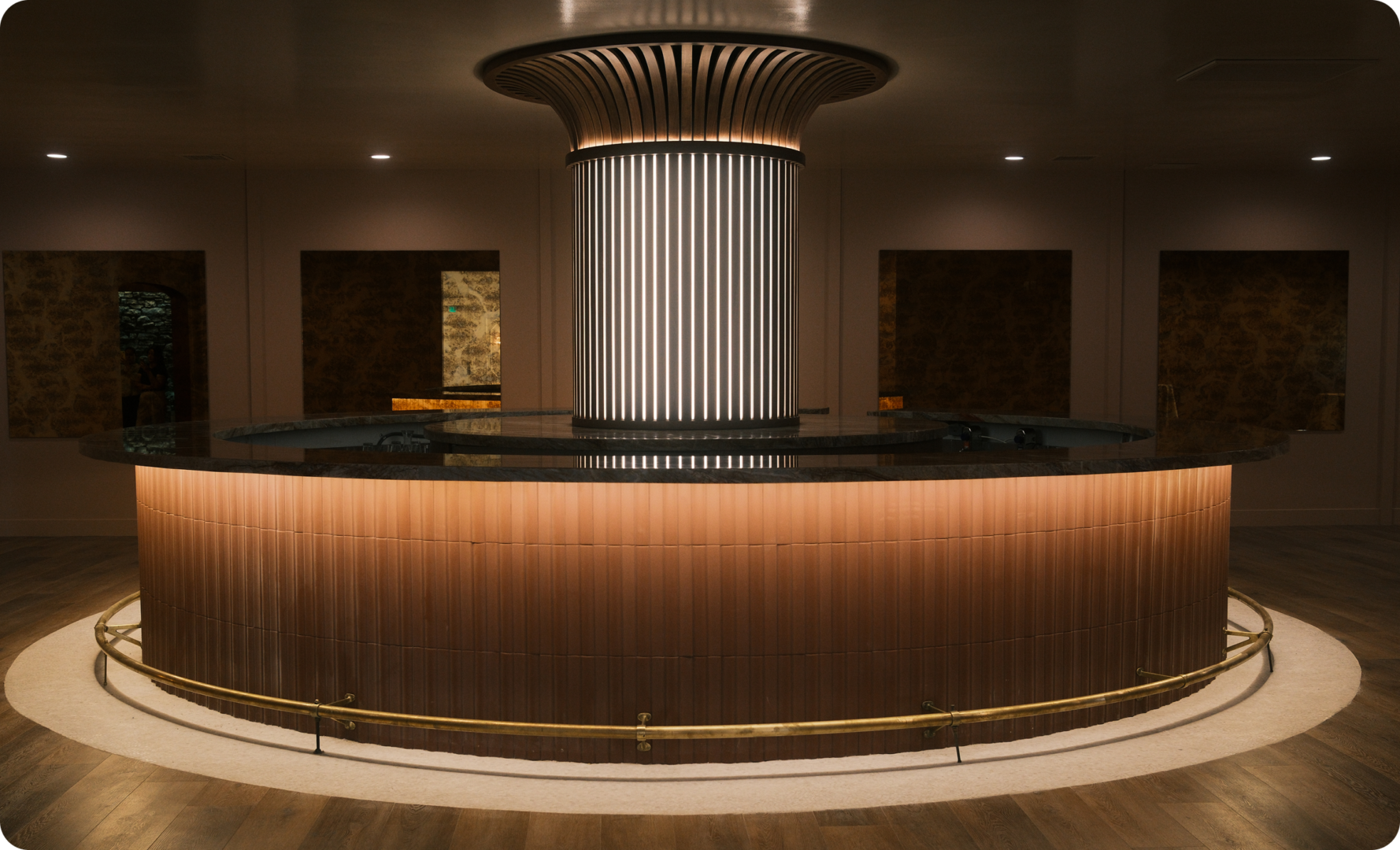 A modern circular bar with a central column featuring vertical lights, surrounded by a curved brass foot rail and wooden flooring, under soft ambient lighting.