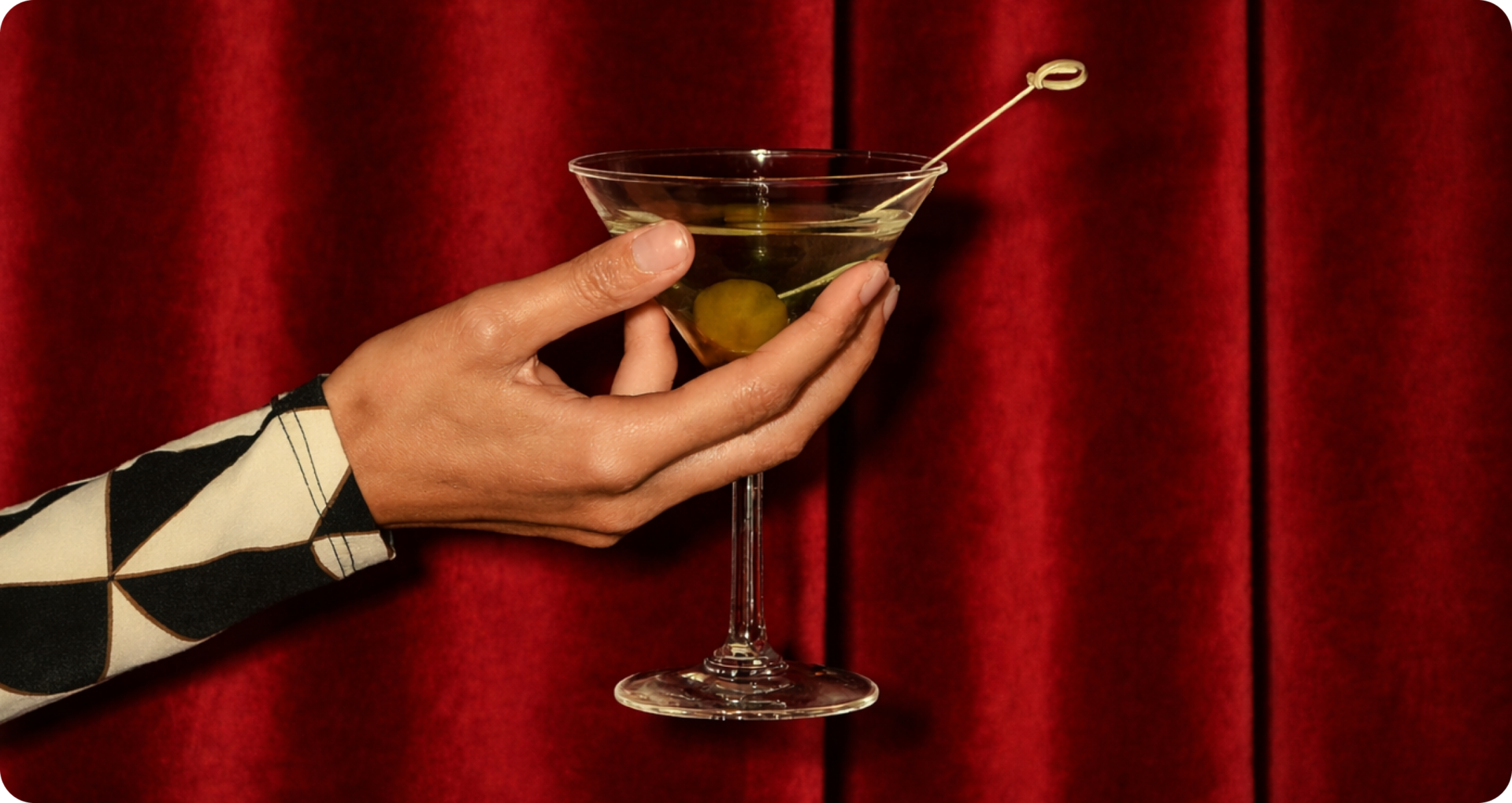 A hand holding a martini glass with an olive garnish, in front of a red curtain. The person wears a patterned sleeve with black and white geometric shapes.
