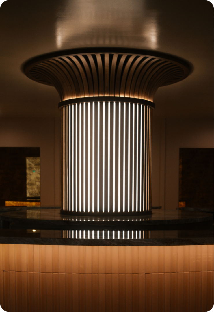 A large, cylindrical light fixture with vertical glowing lines stands in the center of a dimly lit modern room, creating a dramatic, artistic focal point.