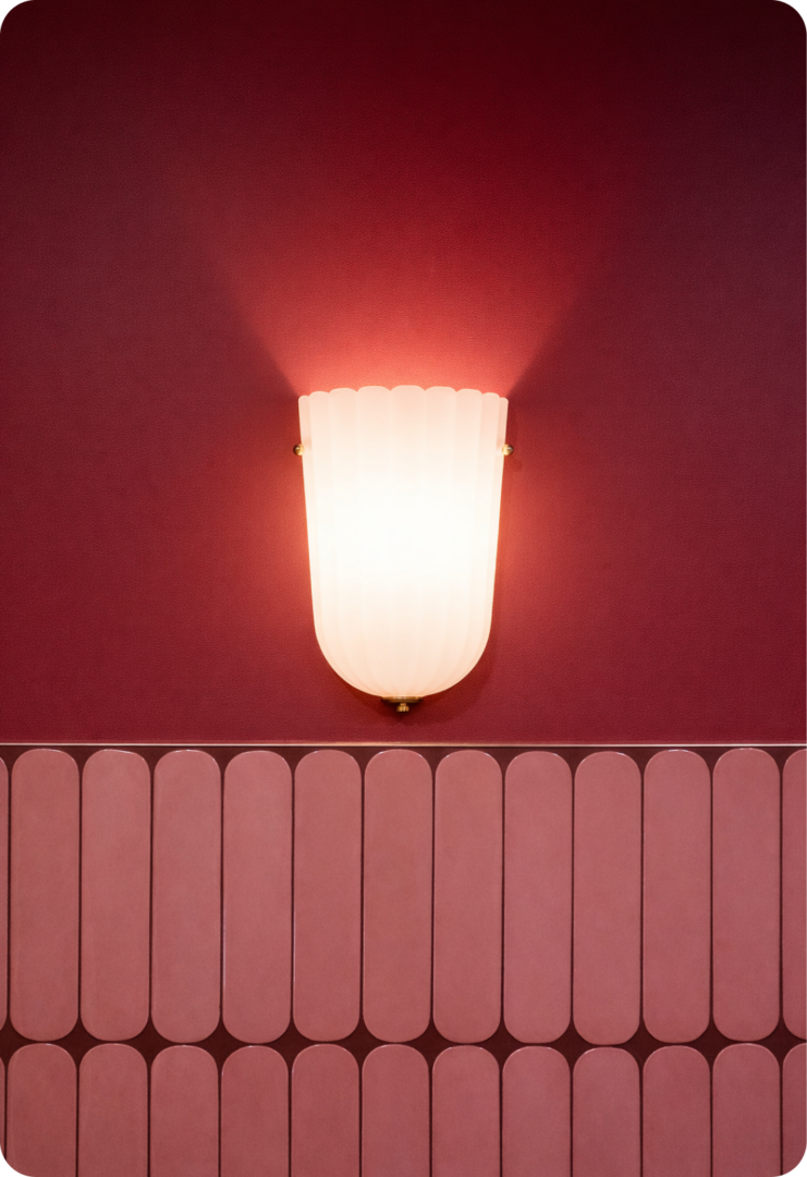 A wall sconce with a frosted, fluted glass shade emits warm light on a deep red wall with pink, rounded rectangular tiles arranged in a horizontal row below.