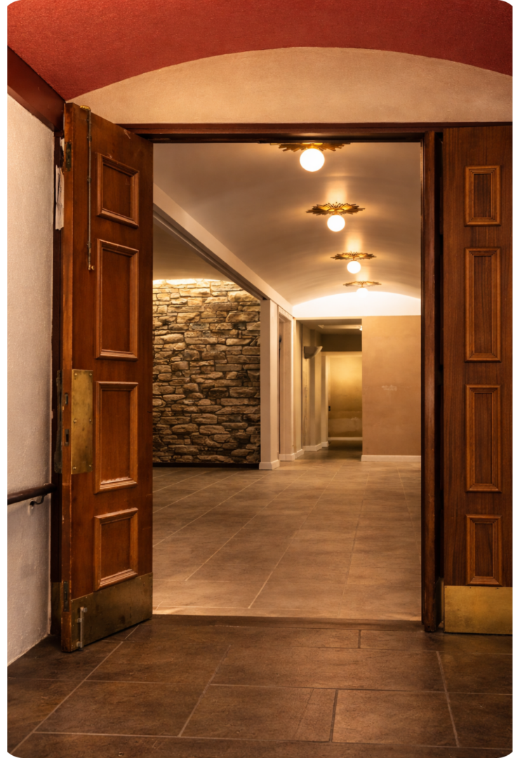 Open double wooden doors reveal a hallway with stone accent wall, modern ceiling lights, brown floor tiles, and neutral-colored walls.