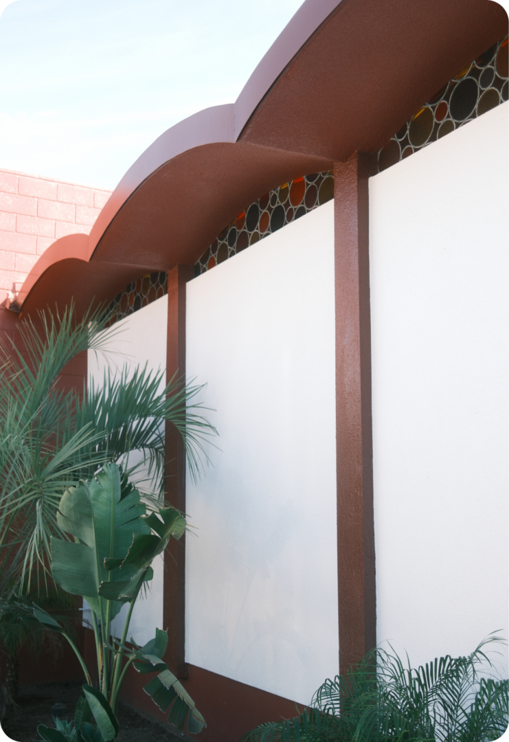 A mid-century modern building facade with white panels, red-brown trim, scalloped roofline, circular window details, and tropical plants in the foreground.