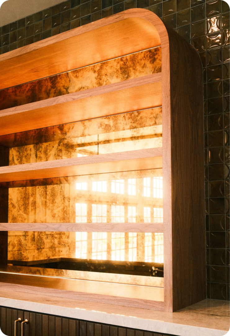 Wooden shelving unit with rounded edges in front of a gold-tiled wall reflecting sunlight and window patterns. The shelves are empty, and the lower part of the image shows a marble countertop.