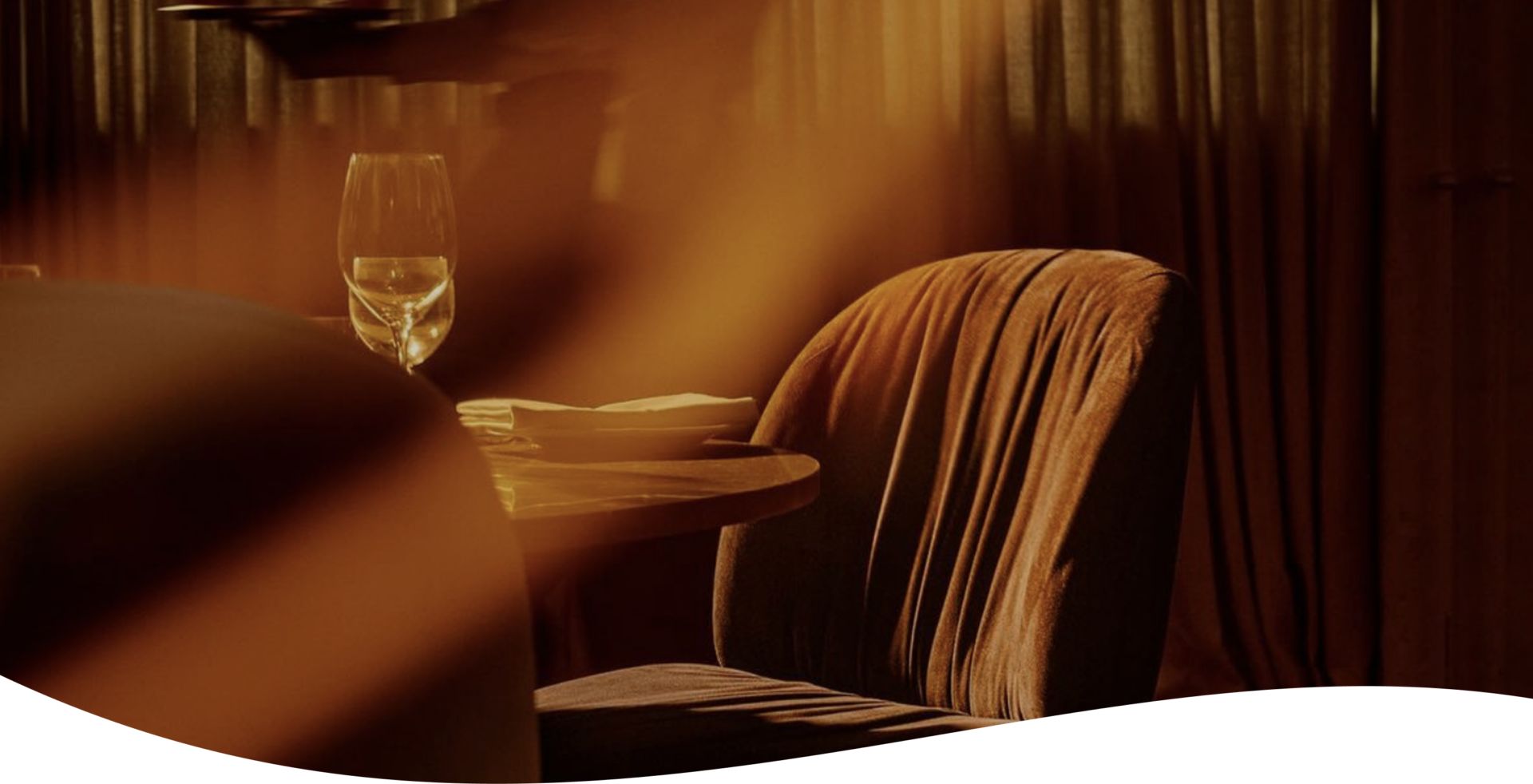 A warmly lit dining area with a glass of water, stacked plates, and a soft, textured chair beside a round table. Sunlight filters through curtains, casting orange-brown shadows across the scene.