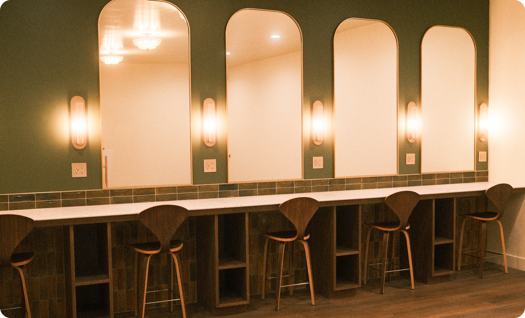 A row of wooden chairs sits at a long counter beneath large arched mirrors and wall sconces, in a stylish, modern room with green walls and tile accents.