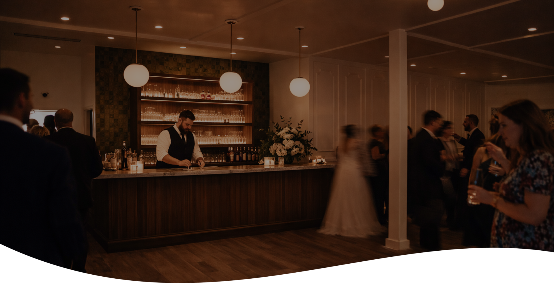 A bartender serves drinks behind a wooden bar in a warmly lit room as guests in formal attire mingle during an event or reception.