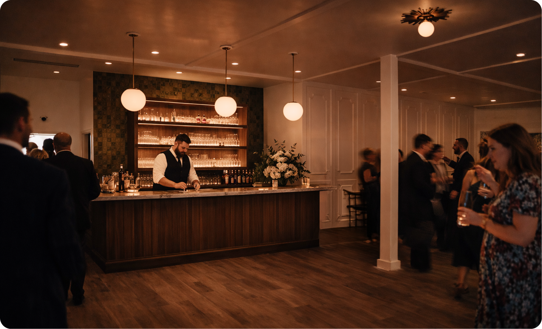 A bartender prepares drinks behind a modern bar, with shelves of glasses and bottles. Elegant guests in formal attire stand and mingle in the softly lit, spacious venue.