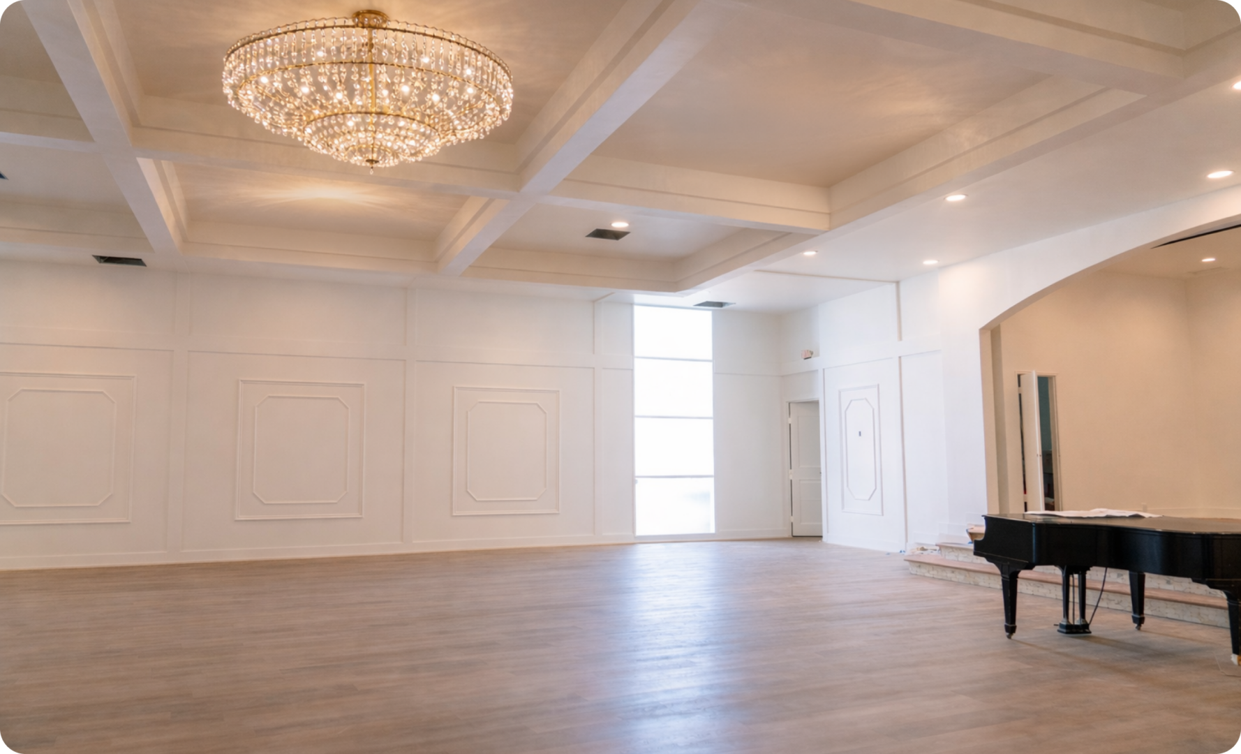 A spacious, elegant room with a chandelier, high ceilings, white walls, wood floors, and a black grand piano near a doorway. Natural light enters through tall windows.