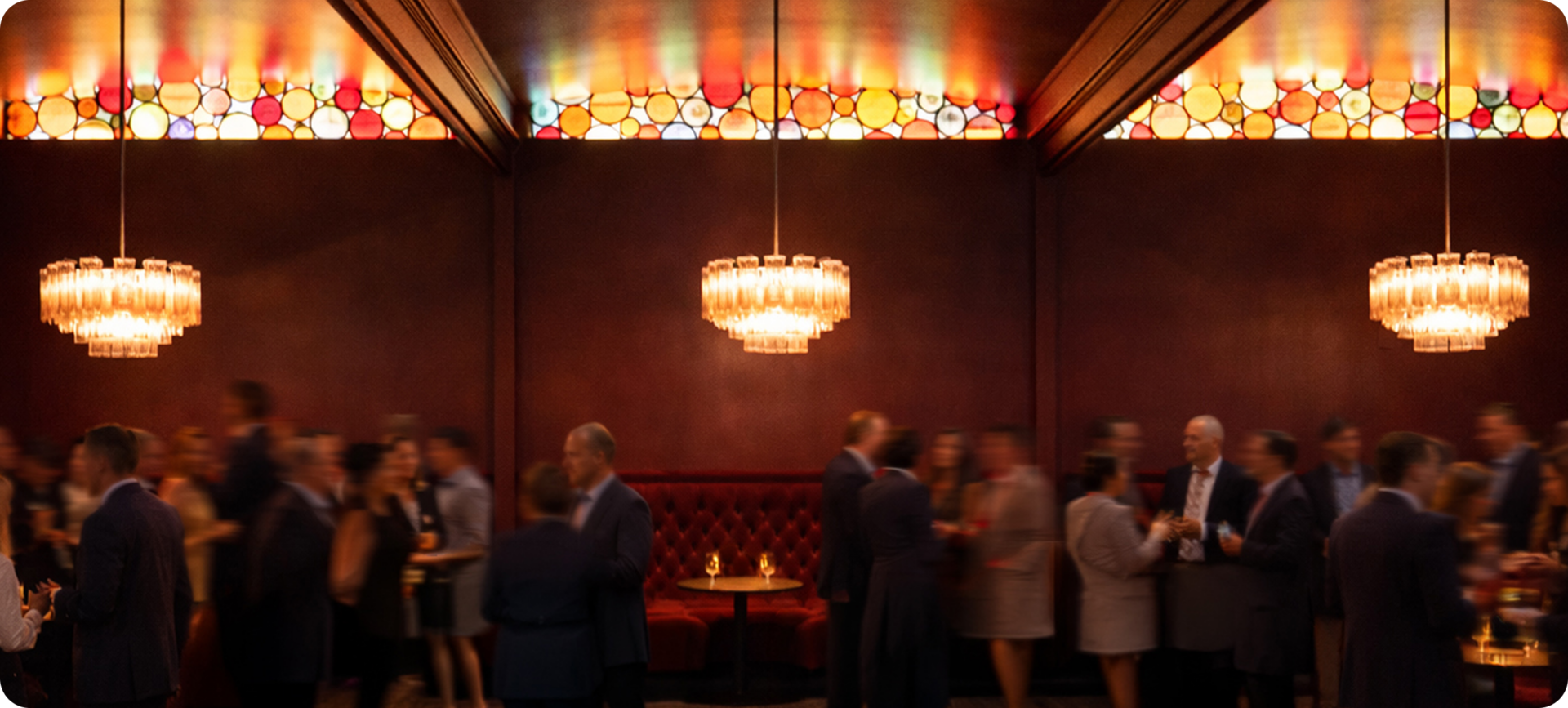 A dimly lit lounge with stained glass ceiling panels, chandeliers, and groups of people in formal attire conversing and holding drinks.