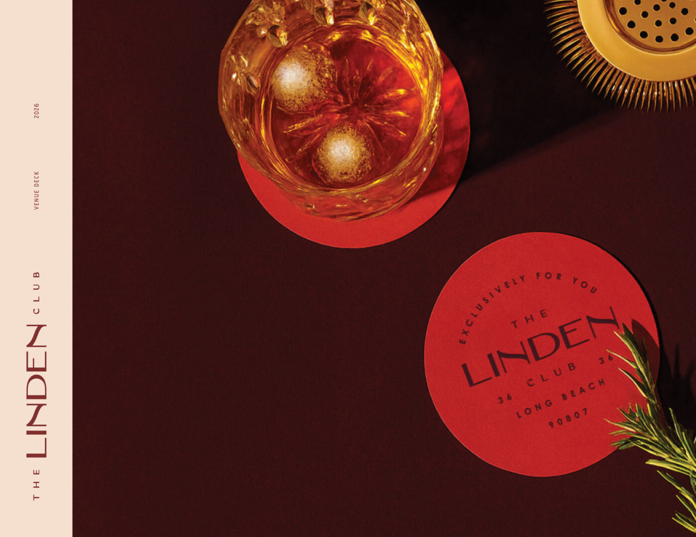 A glass of whiskey with ice sits on a red coaster next to a cocktail strainer and a sprig of rosemary. Another coaster reads The Linden Club, 36 Long Beach #9087 in elegant font.